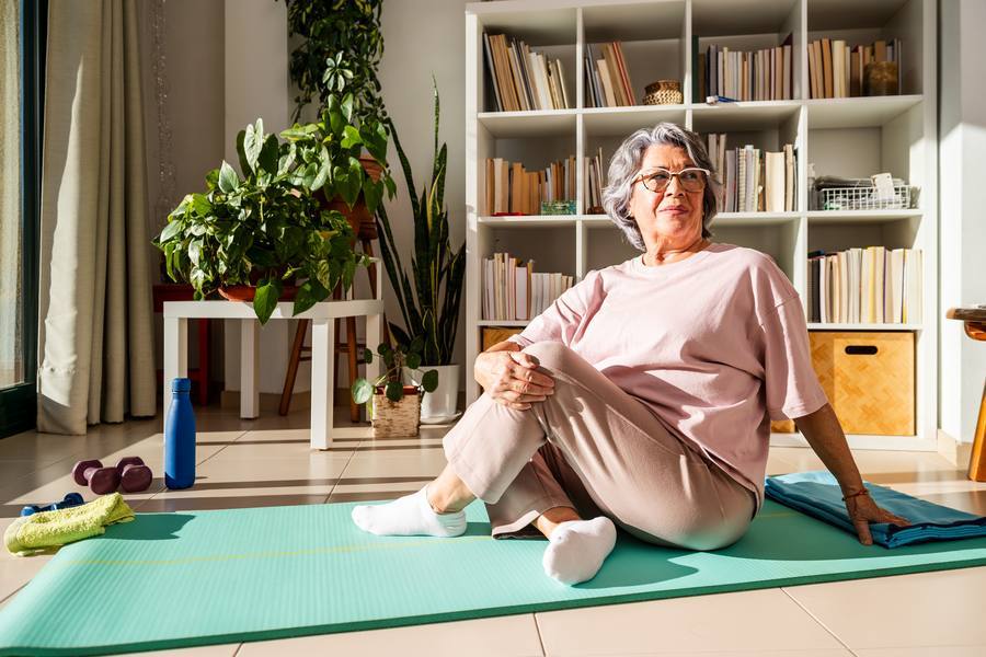 Senior woman sitting on yoga mat at home after stretching workout — fitness after 40
